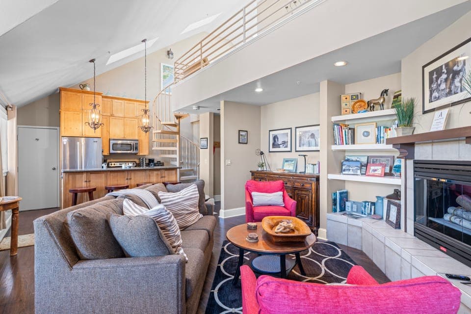 A cozy and eclectic living room with an open kitchen and loft with a spiral staircase, decorated with vibrant pink armchairs. (40B)