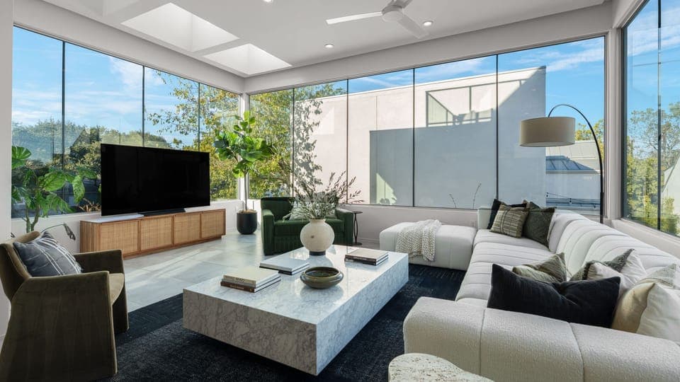 Bright, modern living room with large windows, a light sectional sofa, and a television.