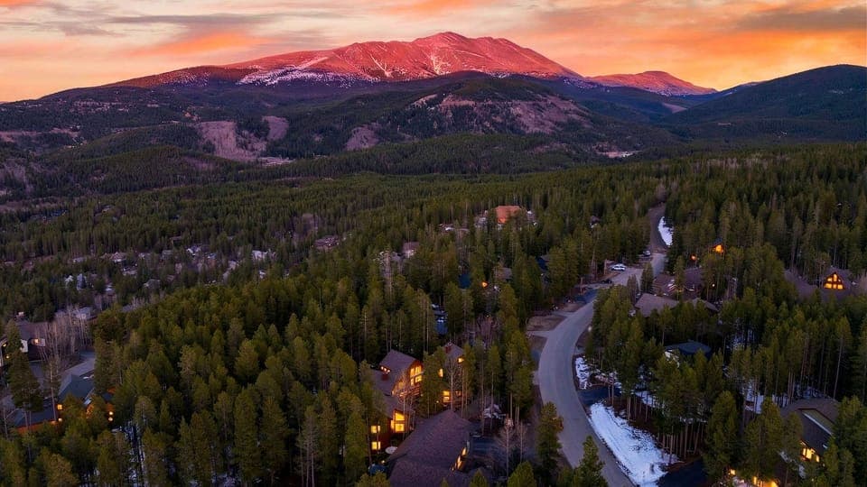Stunning high-altitude shot of the cabin and mountains under a vibrant orange and pink sunset.