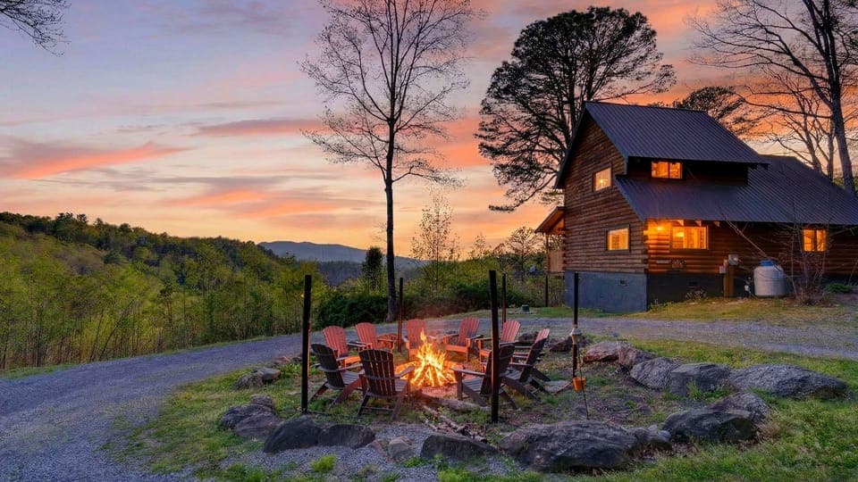Hillside cabin with a glowing fire pit and Adirondack chairs, set against a colorful mountain sunset.