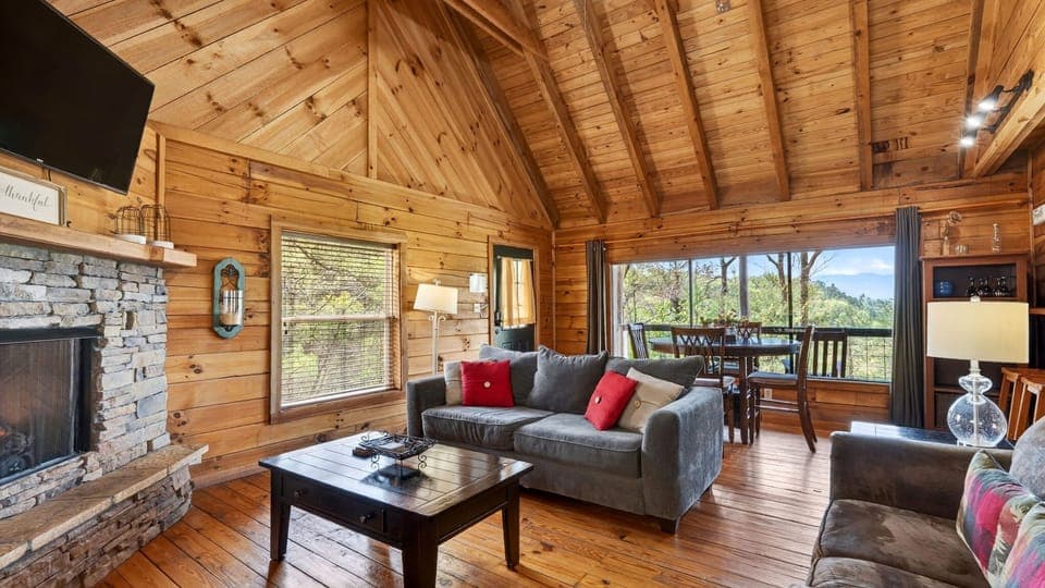 Warm, wood‑lined cabin living room with a stone fireplace, a queen sleeper sofa, soft natural light, and mountain views framed by the dining area windows.