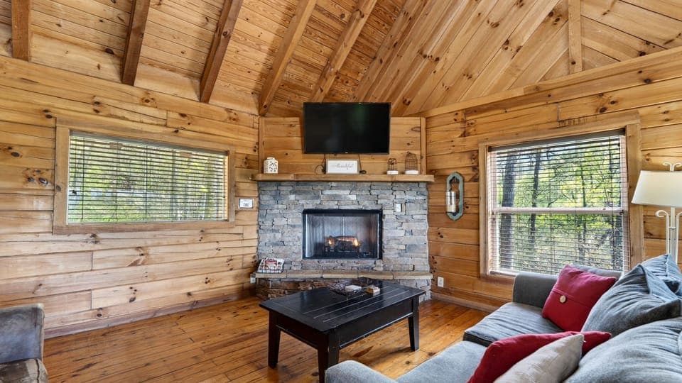 Warm, wood‑lined cabin living room with a stone fireplace, a queen sleeper sofa, soft natural light, and mountain views framed by the dining area windows.