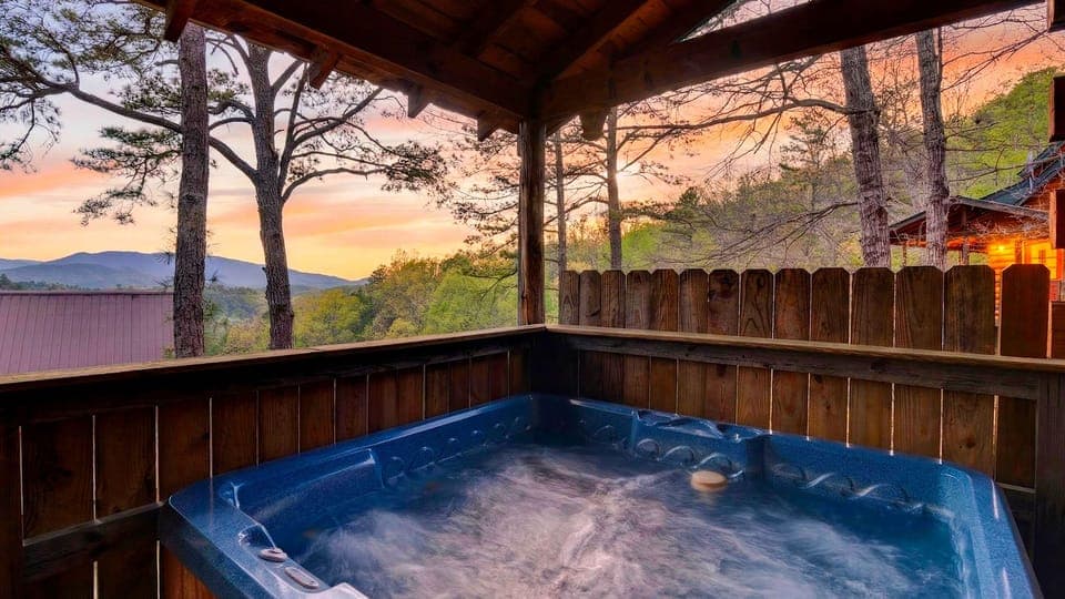 Mountain‑view hot tub retreat with warm cabin glow and a sunset sky painted in pinks and gold.