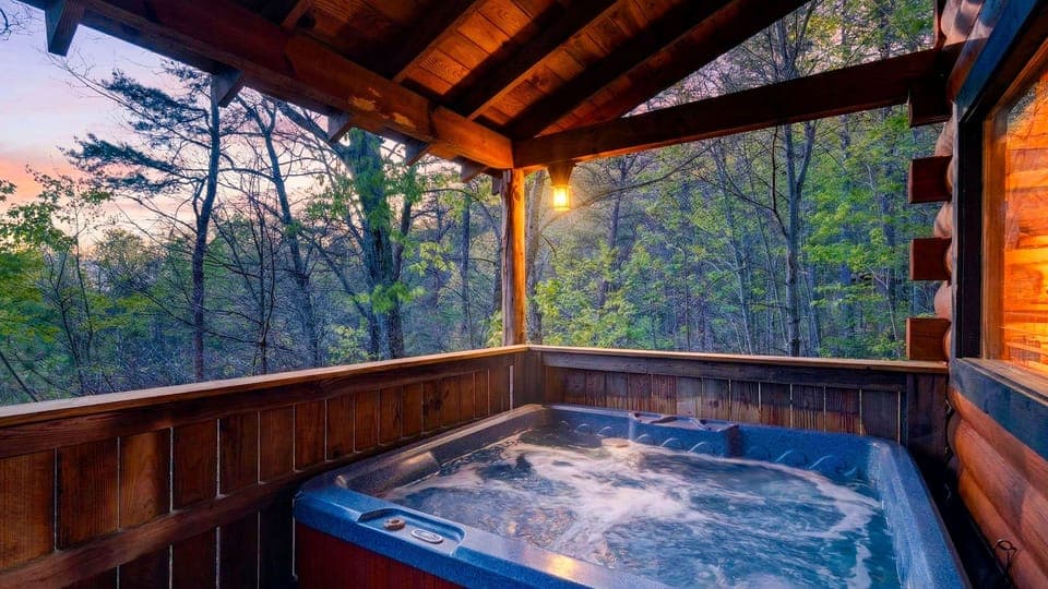 Covered cabin deck with a bubbling hot tub, warm evening light, and peaceful forest views at dusk.