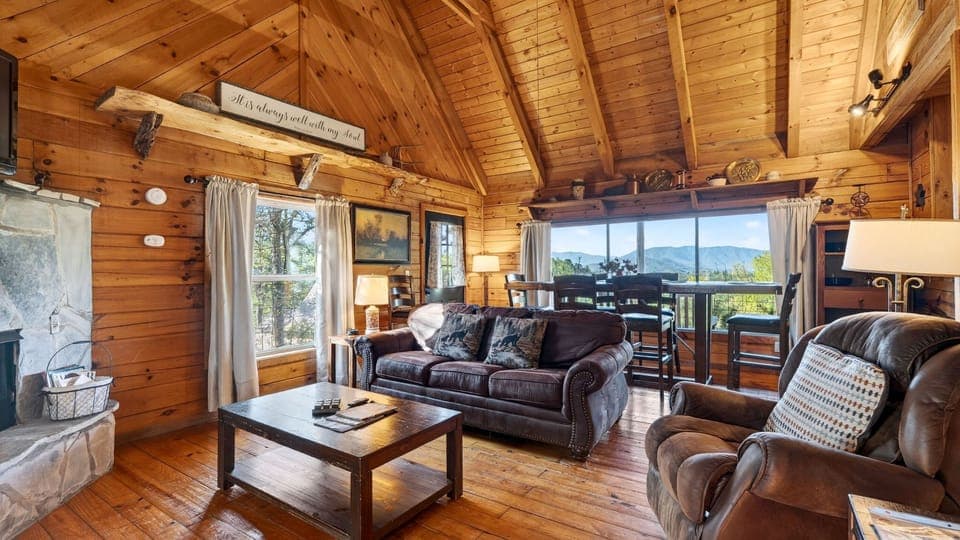 Warm cabin living room with a glowing stone fireplace, a queen sleeper sofa, and sunlight pouring through big mountain‑view windows.