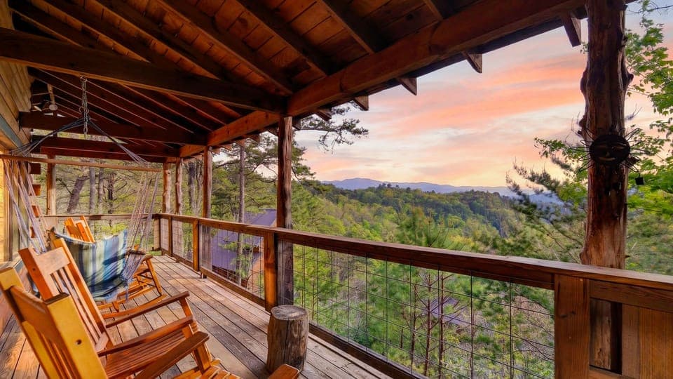 Sunset‑lit cabin deck with rocking chairs and a hammock swing overlooking peaceful forested mountains.