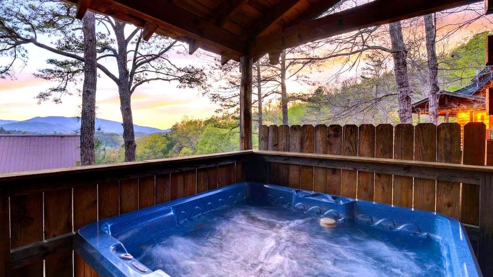 Mountain‑view hot tub retreat with warm cabin glow and a sunset sky painted in pinks and gold. (Elianah's Cabin)