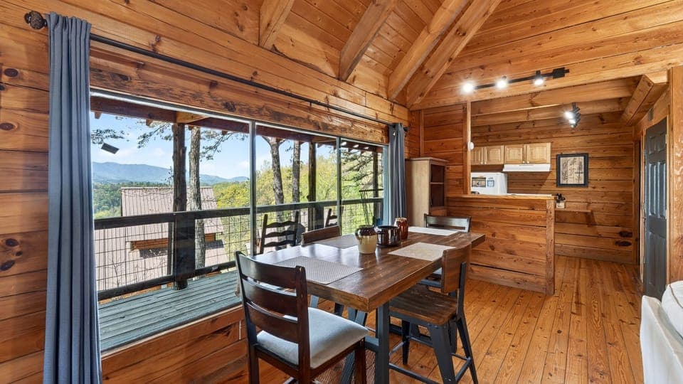 Cozy cabin dining nook with warm wood tones and a picture‑perfect mountain view framed by tall pines. (Elianah's Cabin)