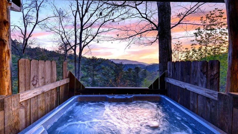 Bubbling hot tub overlooking rolling forested hills under a vibrant sunset sky. (Isaiah's Cabin)