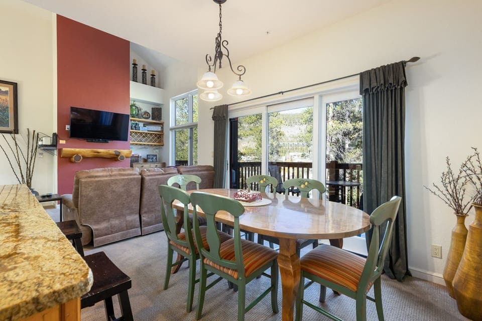 Dining area with a round table and four chairs next to a living room with two sofas facing a wall-mounted TV. A large window with dark curtains looks out onto a forested view.