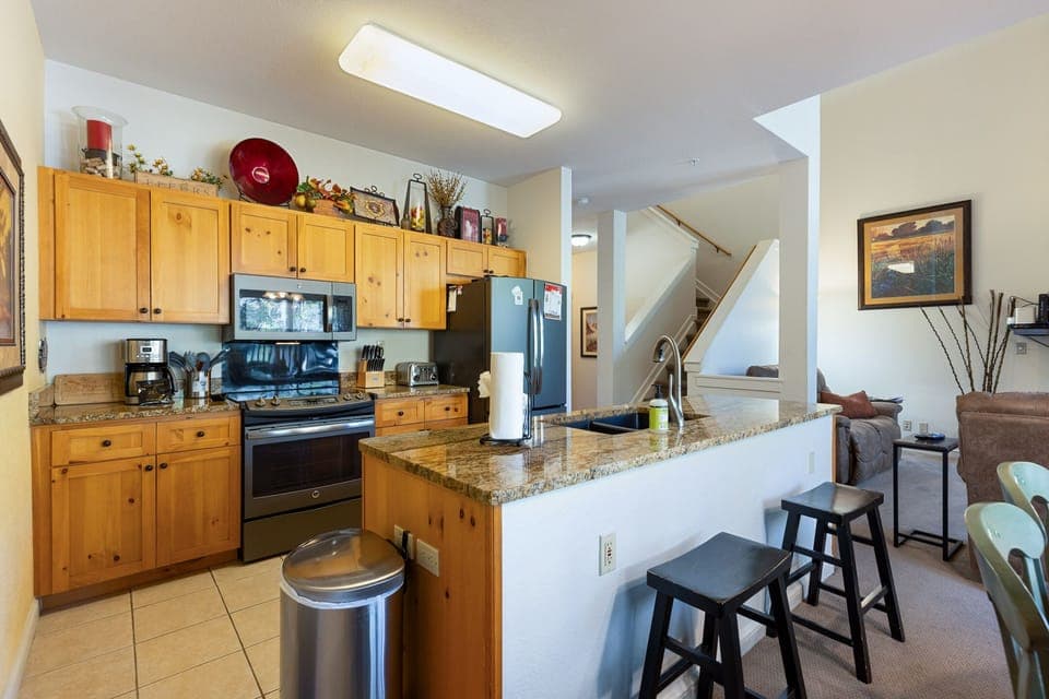 A modern kitchen with wooden cabinets, stainless steel appliances, and granite countertops. A breakfast bar with two stools sits adjacent to the living area and a staircase is visible in the background.