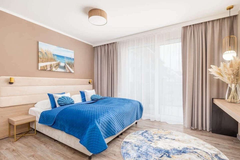 A bright bedroom with a blue bedspread, decorative pillows, and large windows.

