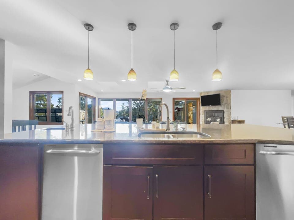 A modern kitchen with a large island sink, pendant lights above, stainless steel dishwasher, and wooden cabinets. Two sets of double doors open to a deck with seating. A fireplace is visible in the background.