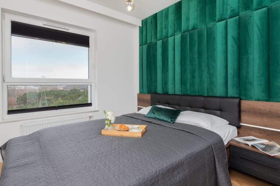 A modern bedroom featuring a luxurious green velvet headboard, a neatly made bed with gray bedding, and a large window allowing natural light.

