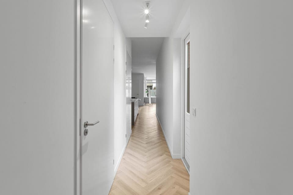 A sleek, minimalistic hallway with crisp white walls, warm herringbone flooring, and soft lighting leading towards an inviting open-concept living space.