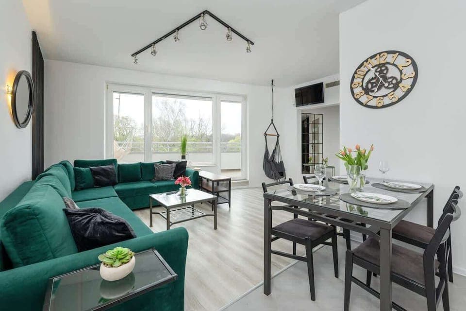 A cozy living room with a round dining table, green velvet chairs, and large windows letting in natural light. Perfect for relaxed meals or morning coffee.