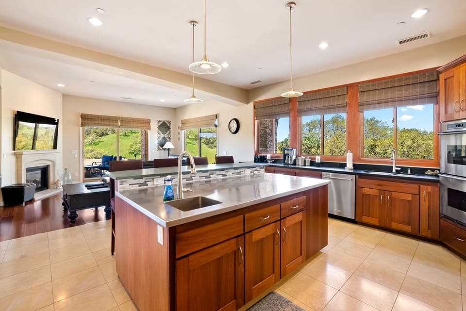 This expansive gourmet kitchen features a massive stainless steel island with a prep sink and custom wood cabinetry. Large windows frame stunning views of the rolling green hills, seamlessly connecting the interior to the breathtaking landscape.