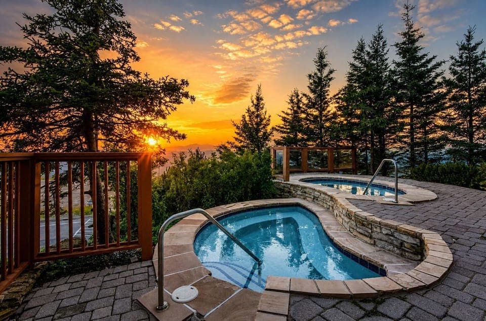 Two stone-bordered hot tubs sit on a paved patio surrounded by tall evergreens, glowing in sunset light.