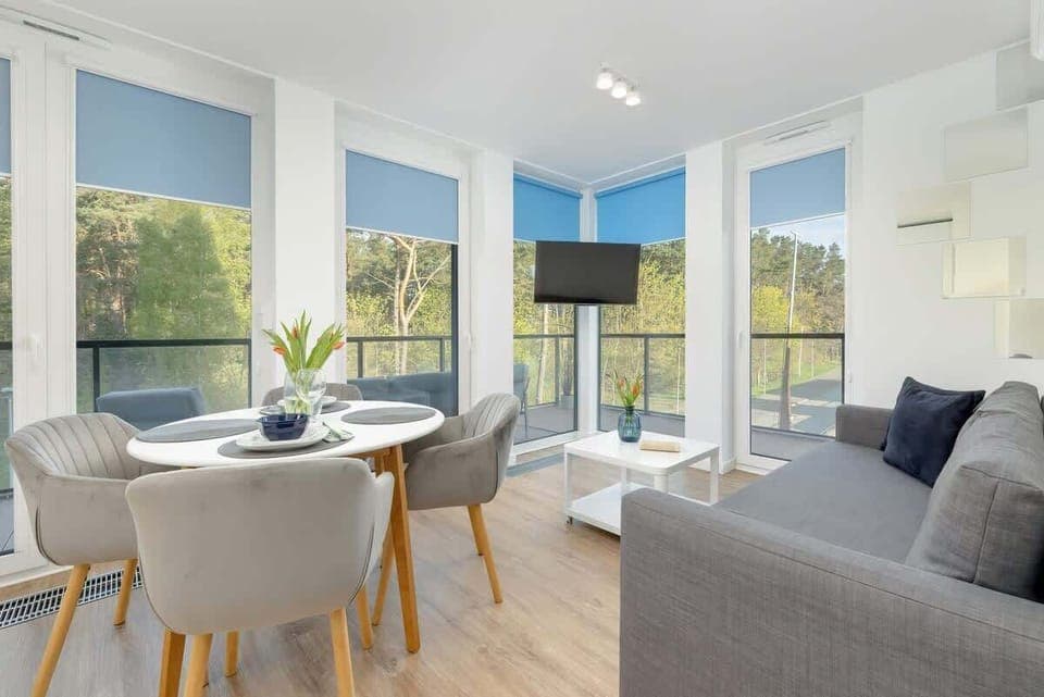 A bright living room with large windows, a round dining table, and modern decor. The room has light wood flooring and a cozy atmosphere.