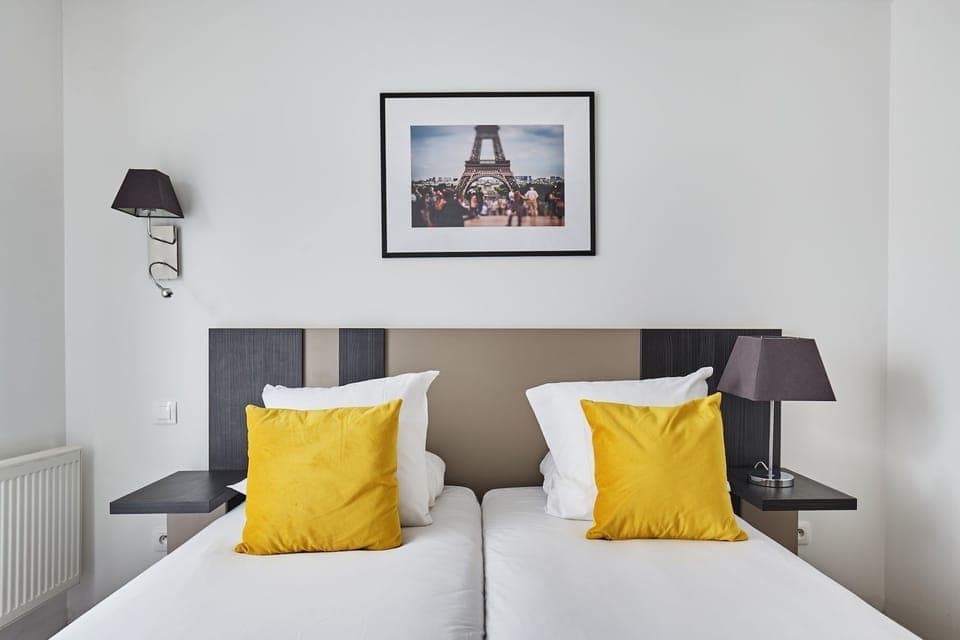This cozy studio features zip beds adorned with bright yellow pillows, complemented by modern wall sconces and a framed photograph of the Eiffel Tower.