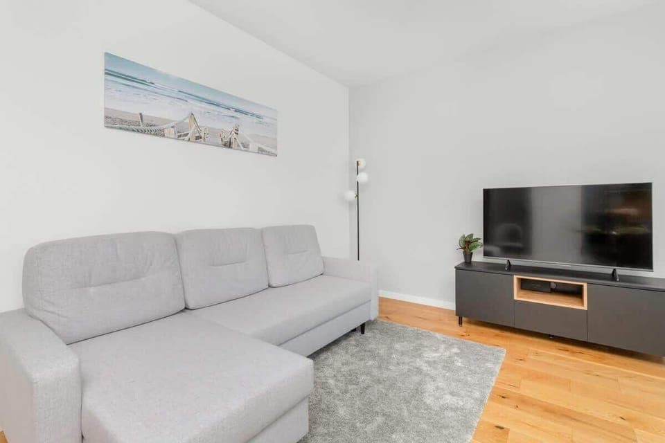 A modern living room featuring a dining area, with a light gray sofa, a TV on a console, and wooden flooring.