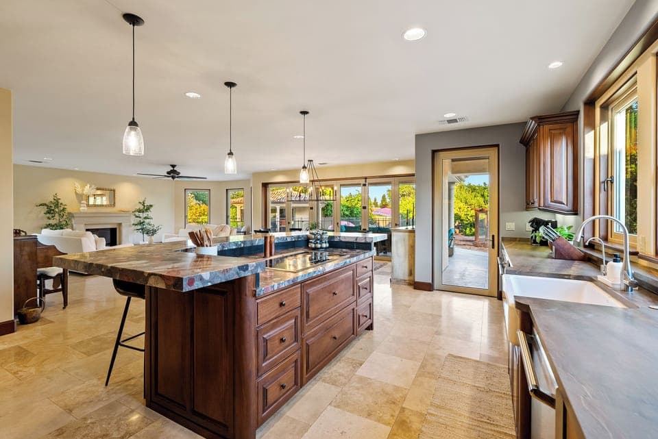 This grand gourmet kitchen and open-concept living area offer a massive stone-topped island with a built-in cooktop, elegant wood cabinetry, and a farmhouse-style sink. 
