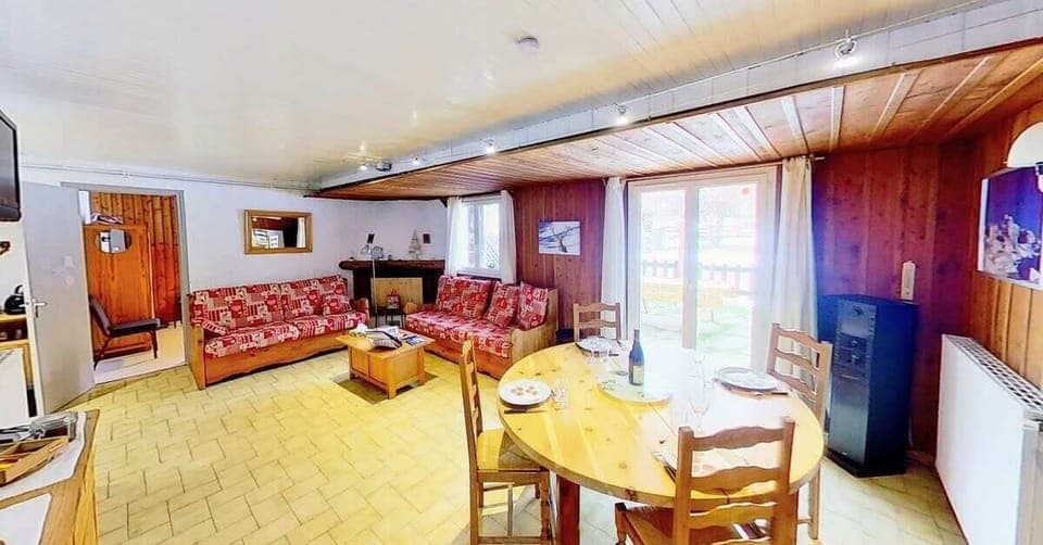 The spacious living area features a comfortable sofa with colorful patterned cushions, a wooden coffee table, and a dining table with chairs set for four. The large window provides a view of the outdoor terrace and surrounding natural landscape. The woode