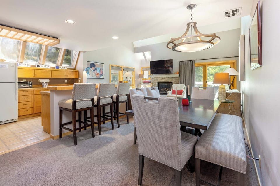 A modern dining and kitchen area with a wooden dining table, upholstered chairs, a kitchen island with bar stools, stainless steel appliances, a fireplace, and a wall-mounted TV.