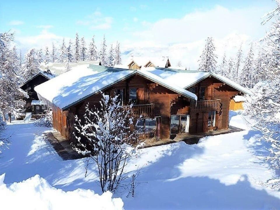 The chalet is a charming wooden structure with a snow-covered roof, situated amidst a picturesque winter landscape. The surrounding trees are laden with snow, creating a serene and idyllic ambiance. This charming chalet is perfect for those seeking a cozy