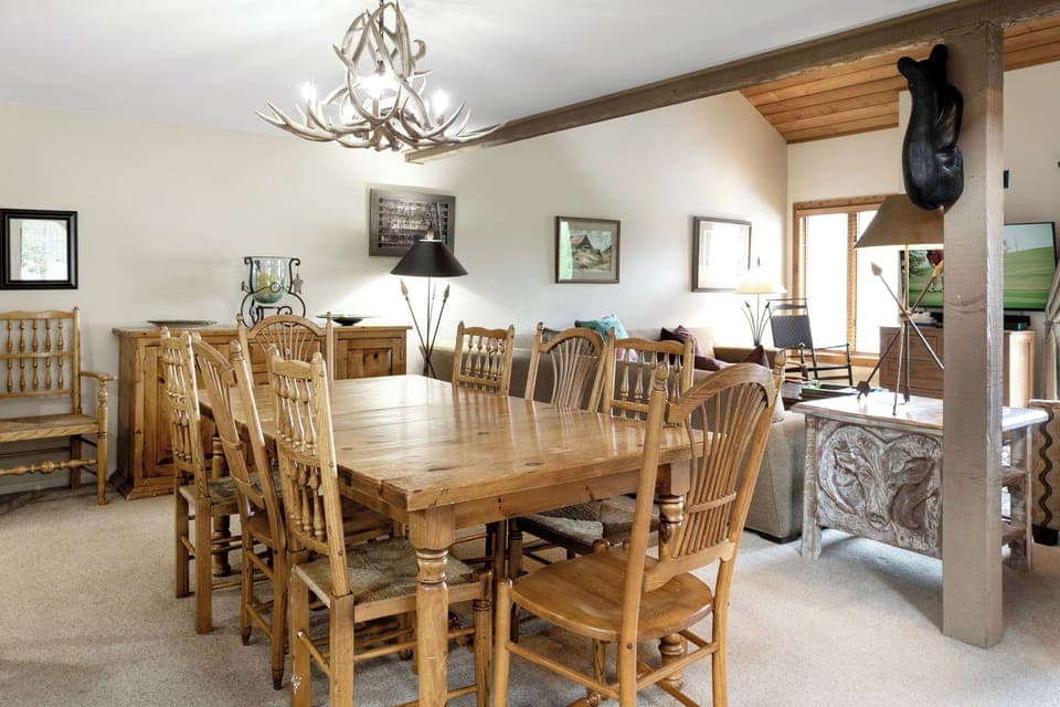 A rustic dining room features a wooden table with eight chairs, an antler chandelier, and assorted decor. The adjacent living area includes a sofa, armchairs, and a TV.