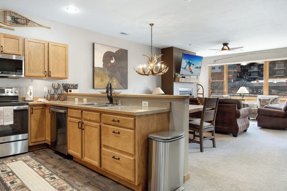 Open-concept kitchen and living area with wooden cabinets, stainless steel appliances, antler chandelier, and large windows. Decor includes animal art and ski-themed picture.