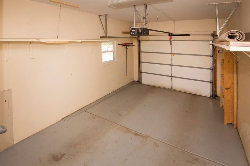 An empty, clean garage interior with beige walls, a small window, a rolled-up carpet in the corner, and a closed garage door with an automatic opener.