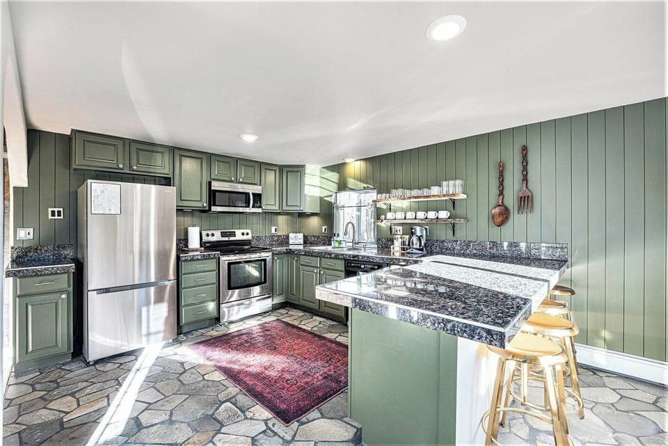- Cook in style with sage green cabinetry, granite counters, and stainless appliances
- Charming open shelves hold a full coffee bar and everything you need to dine in
- Stone floor, oversized utensils, and Persian style rug add rustic elegance