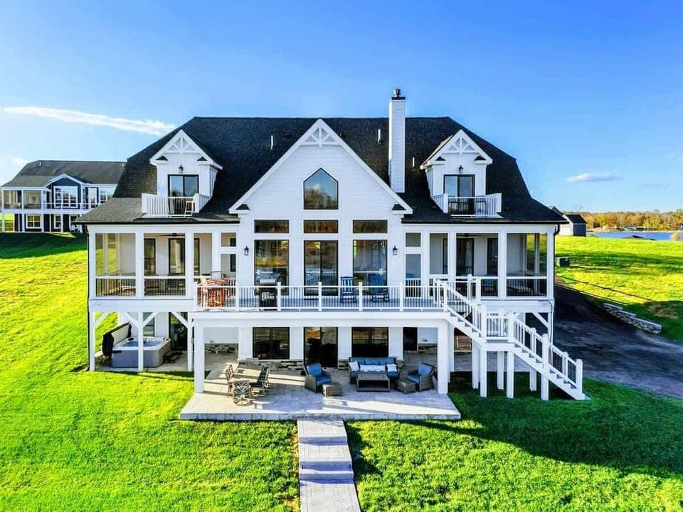 Expansive lakeside home with a modern farmhouse design, multiple balconies, screened porches, and a spacious outdoor patio for relaxation and entertainment.