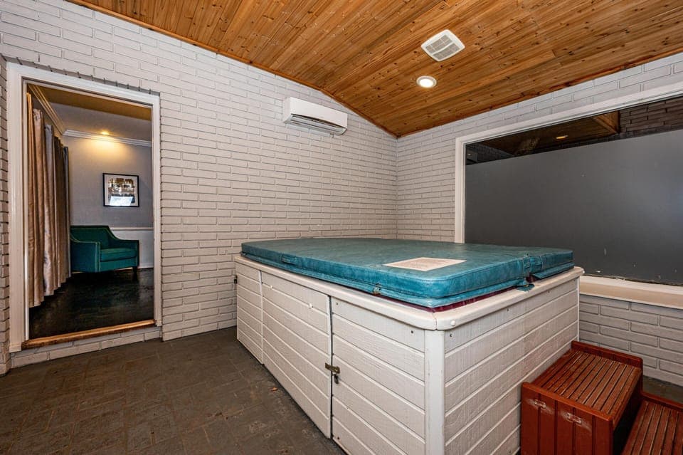 Hot tub room with white brick walls, wood-paneled ceiling, frosted window, and teal cover on tub.