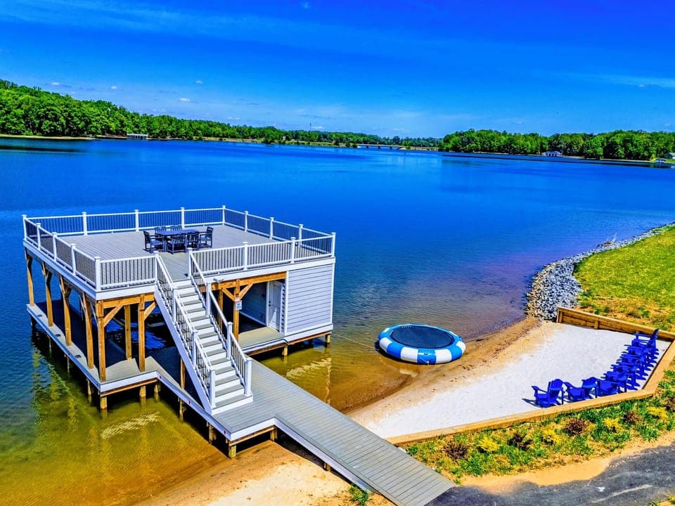 Backyard & Beach - The lakefront layout from above: two-story dock, floating water trampoline, sandy beach lined with Adirondack chairs.