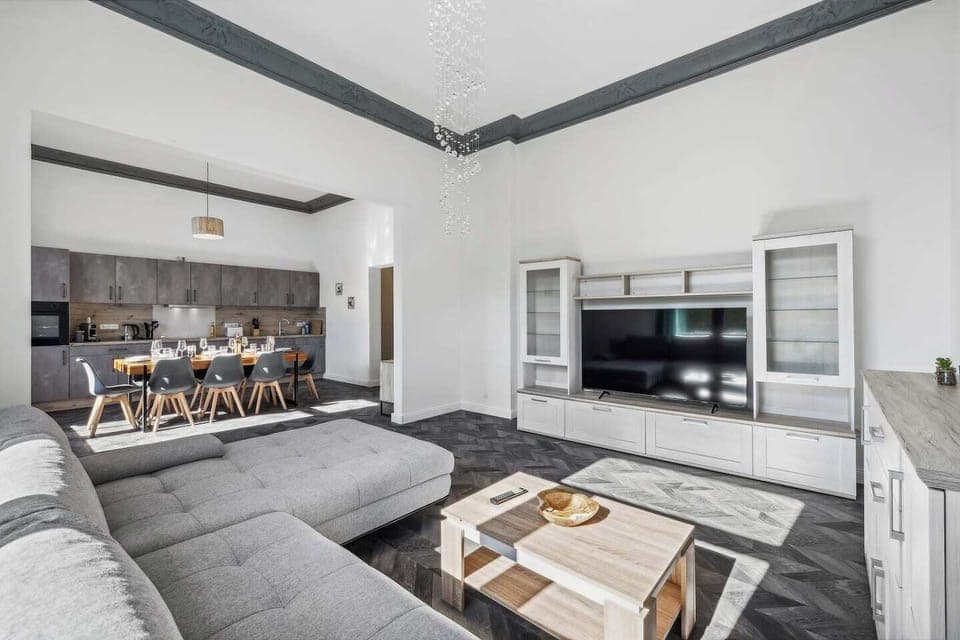 Helles Wohnzimmer mit großer grauer Couch, modernem TV-Möbel mit Flachbildfernseher sowie offener Verbindung zur Küche mit Essbereich.