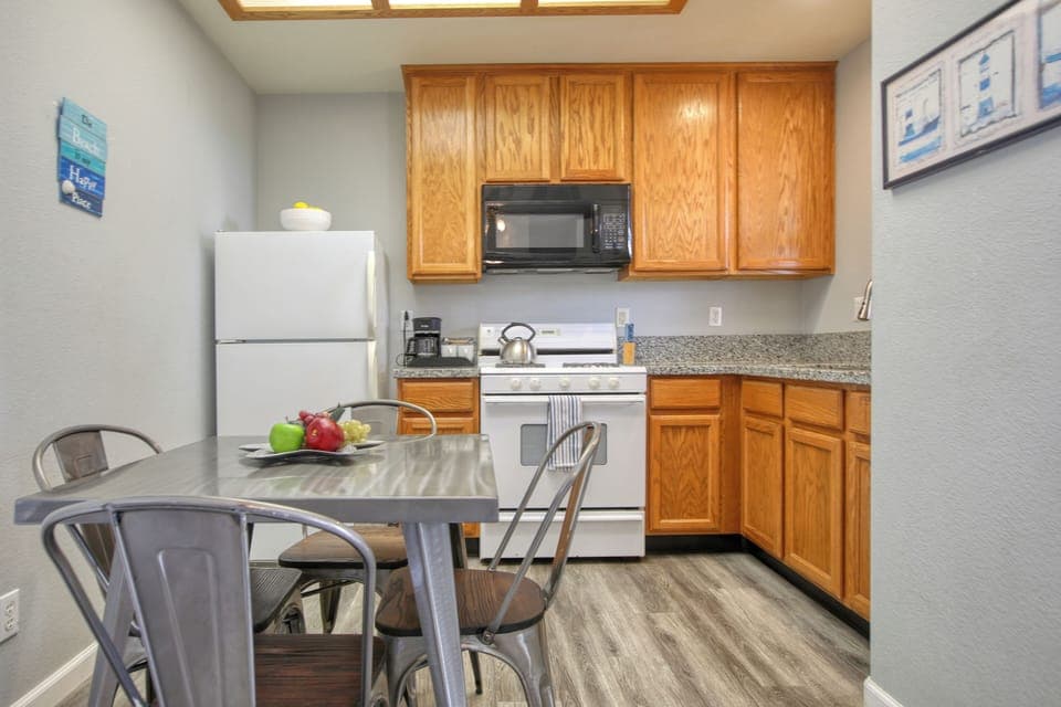 This cozy kitchen features warm wooden cabinetry, a compact dining area with metal chairs, and modern appliances, all set against stylish gray-toned flooring.