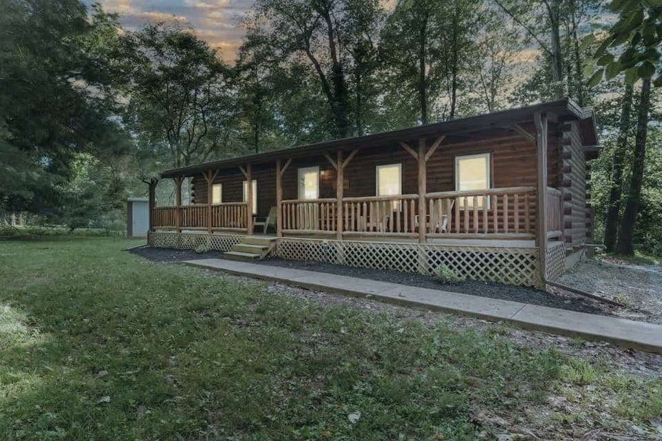 Rustic log cabin surrounded by lush trees, featuring a welcoming front porch with seating and warm glowing windows at dusk, perfect for a cozy retreat in nature ✨