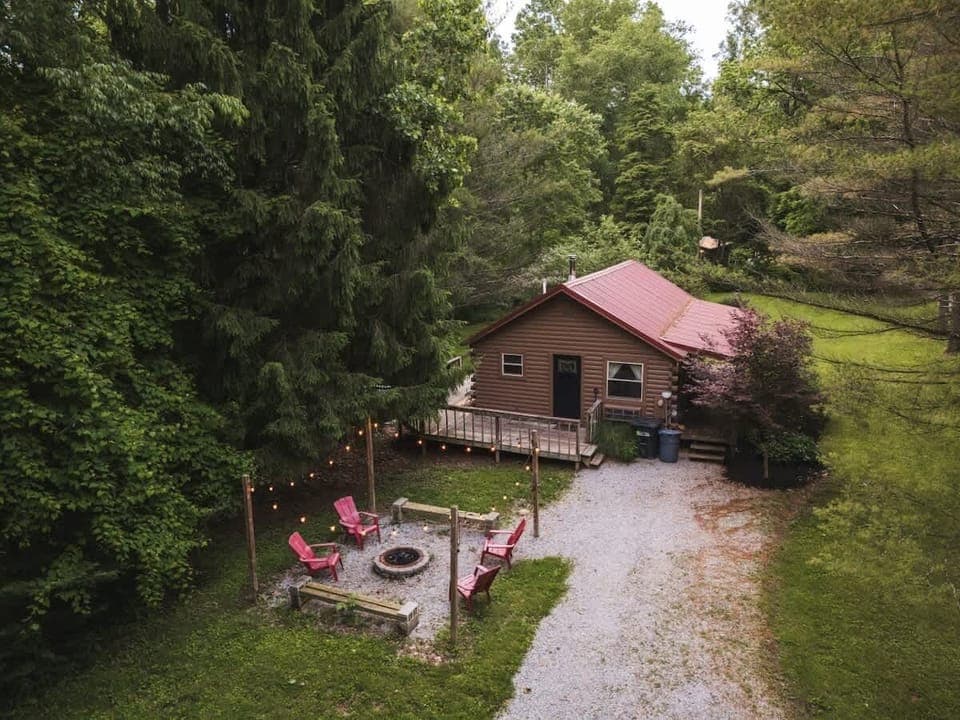 Charming forest cabin with a cozy fire pit surrounded by red chairs, perfect for evening gatherings. A serene natural retreat with lush greenery and warm outdoor lights to set the mood ✨