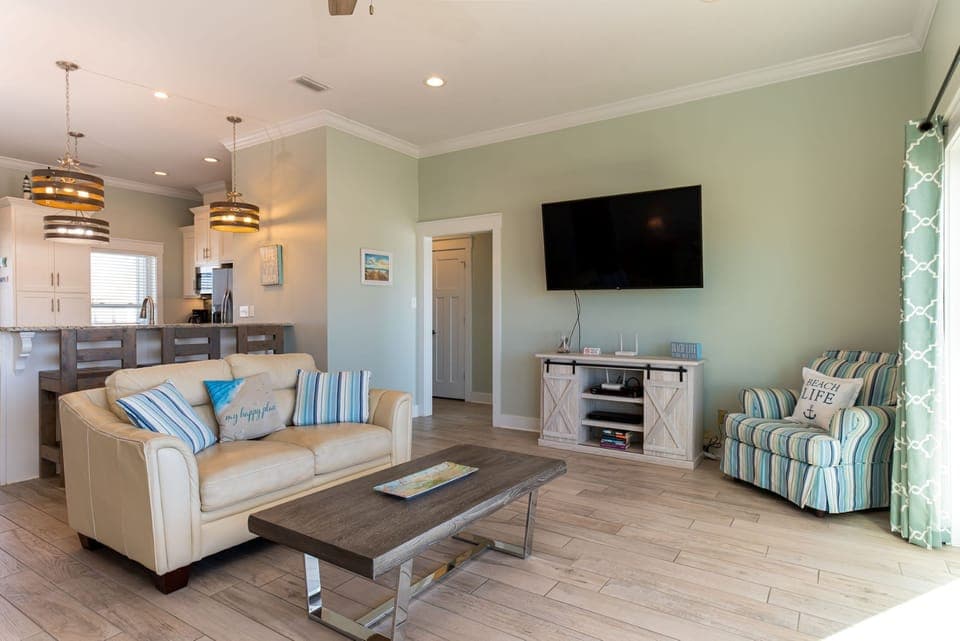 Unwind in your bright, coastal-themed living space featuring comfortable seating, large TV, and open connection to the modern kitchen.