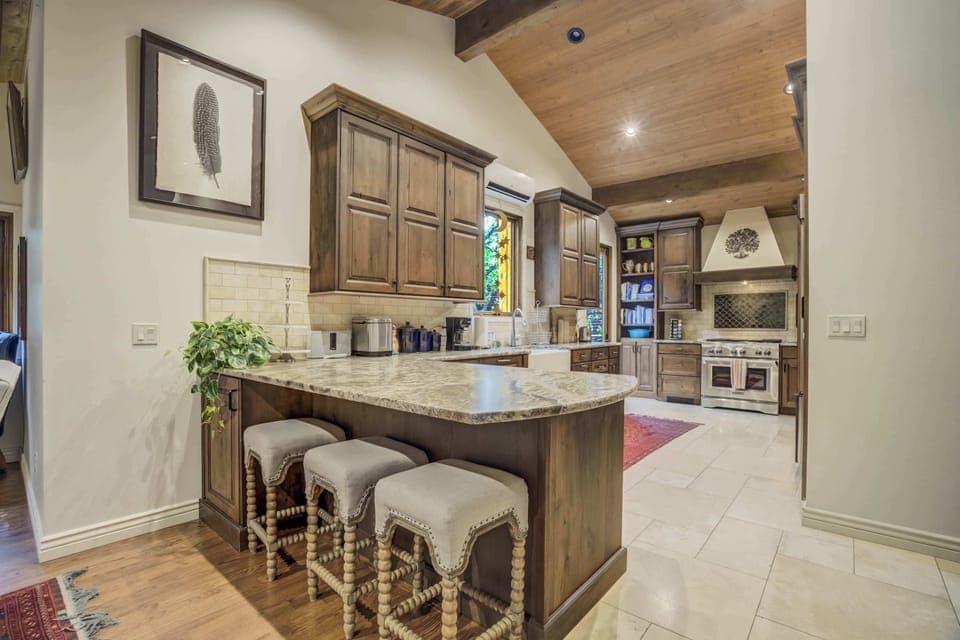 A wide kitchen island with cushioned stools invites casual breakfasts and conversation, surrounded by wood cabinetry and warm lighting—an easy space to prep meals, pour drinks, and linger while dinner comes together.