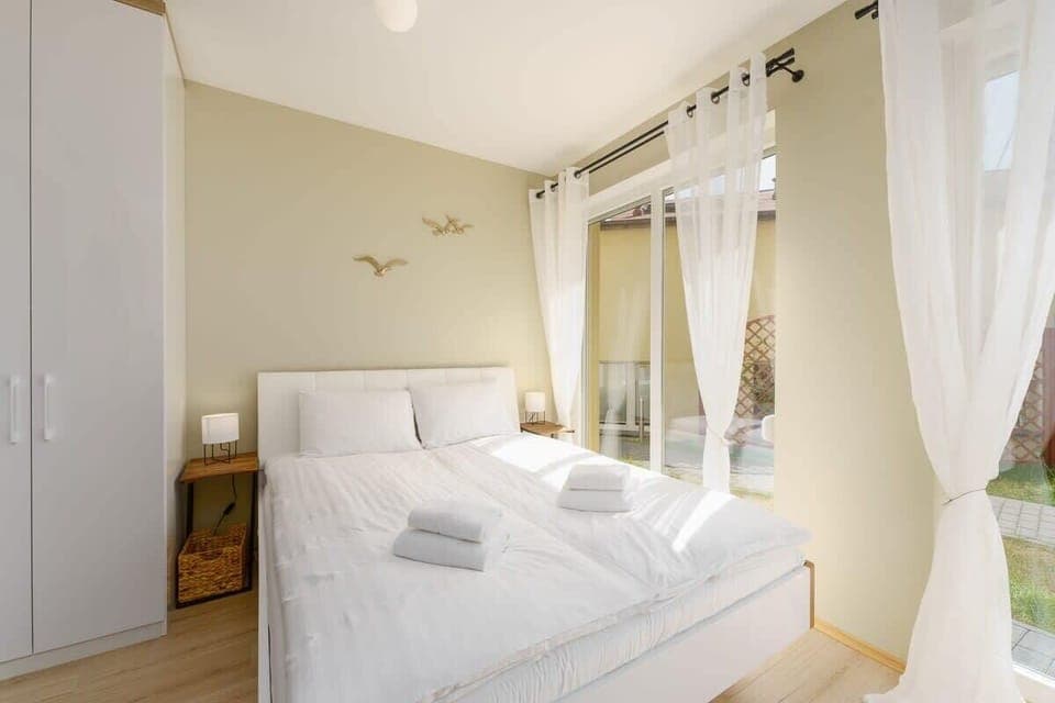 A bright bedroom with a large bed, soft white bedding, and floor-to-ceiling windows. The room has a light, airy atmosphere with plenty of natural light.
