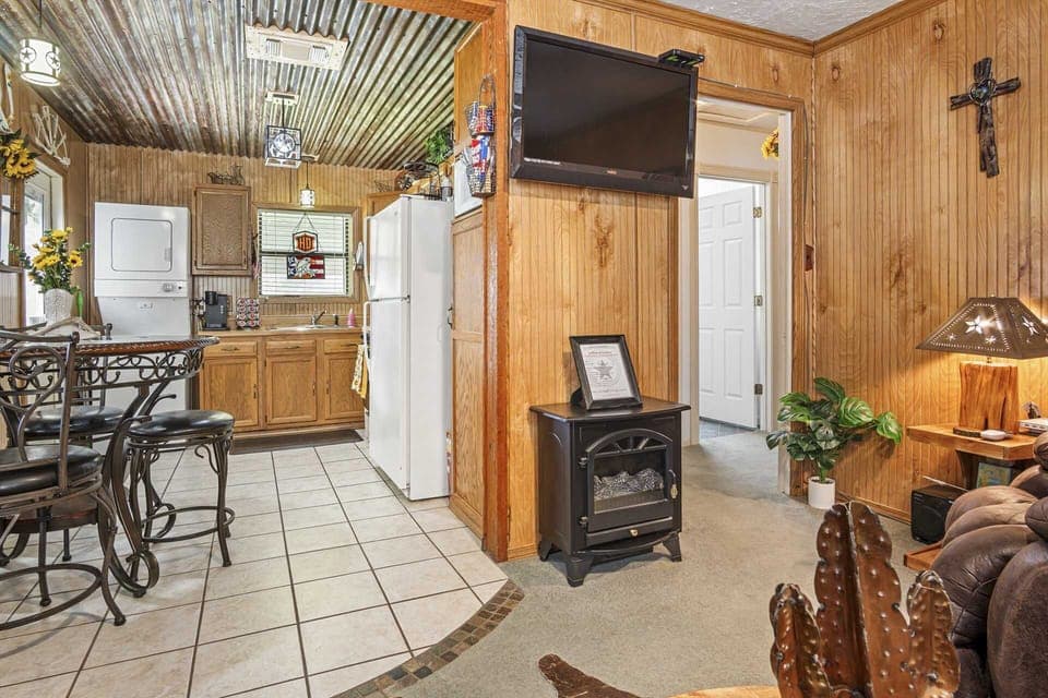 A country-style living area seamlessly transitions into a compact kitchen, featuring wood-paneled walls, a wall-mounted TV, and an electric fireplace, creating a cozy and functional space.