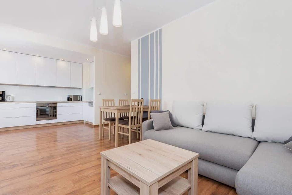 Corner sofa with sleeping function and soft cushions at which there is a quart table. Next to it is a dining table for four. In the background you can see the kitchenette with white furniture. 