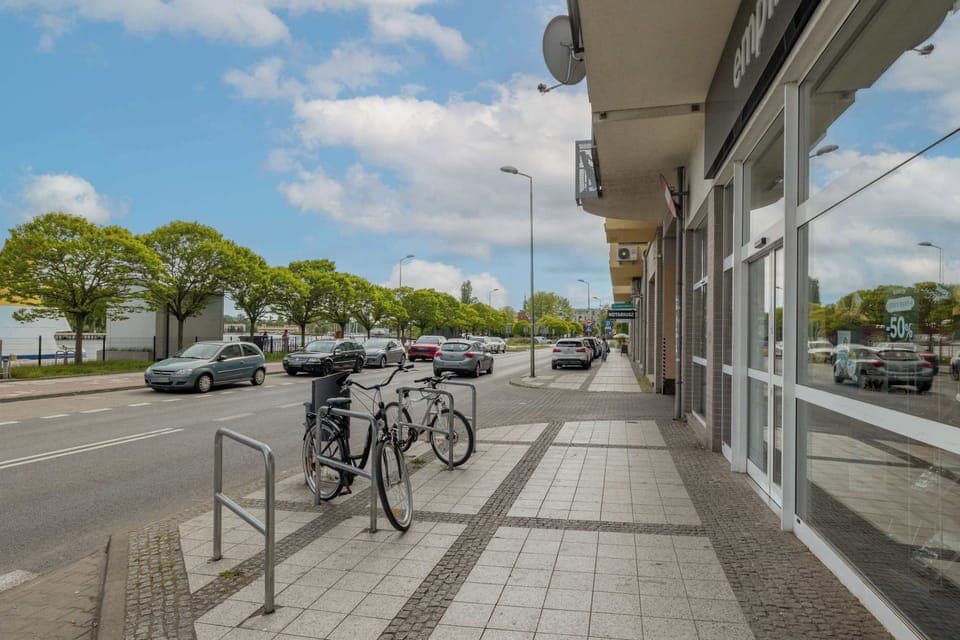 A view of the street with parked bikes, trees lining the path, and apartment buildings in the background.

