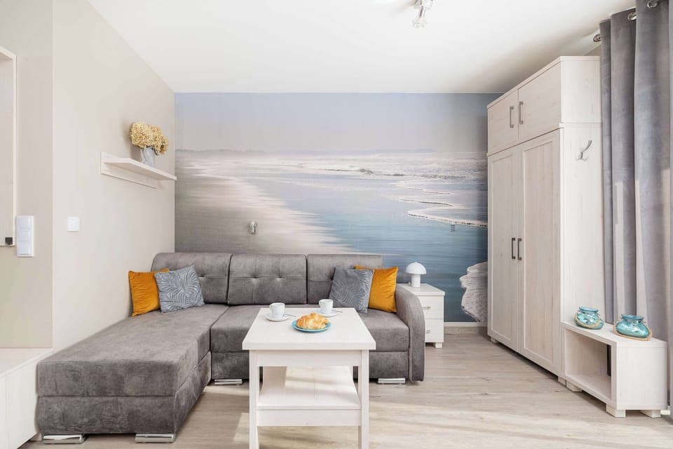 Modern living room with a sectional sofa, gray and orange pillows, and a large scenic wall mural depicting a seaside view. There is a coffee table and large windows letting in plenty of natural light.