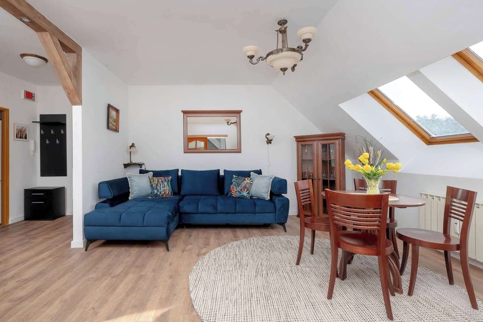 A spacious living room with a blue sofa, wooden flooring, and slanted ceilings, creating a cozy attic-style ambiance. A dining table and chairs are placed in the open-plan space.