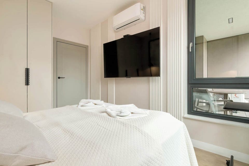 A comfortable bedroom with a wall-mounted TV, air conditioning, and a desk. The space blends sleep and functionality in one.