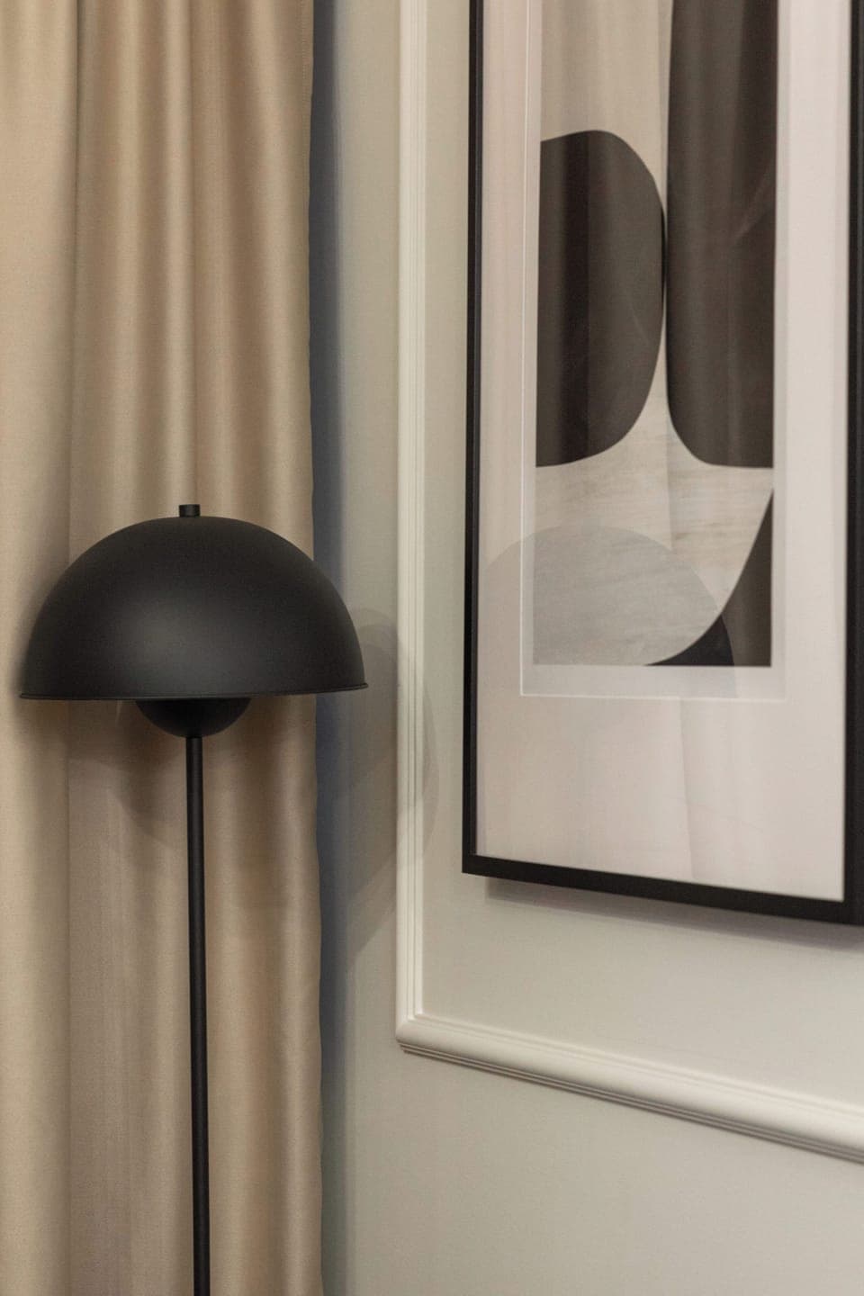 A close-up detail of a beige curtain next to a black wall lamp and a black-and-white framed poster. The soft fabric contrasts with the bold fixture and artwork.

